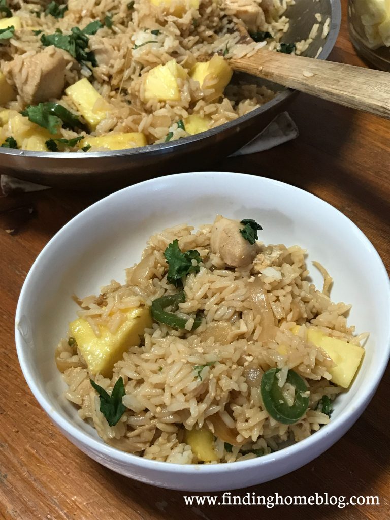 A close up shot of a bowl with fried rice, jalapeños, pineapple, and cilantro; a large skillet holding more of the same in the background.