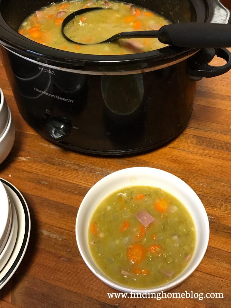 A crockpot full of split pea soup with a ladle sticking out of it. A bowl of the soup is dished out in the foreground