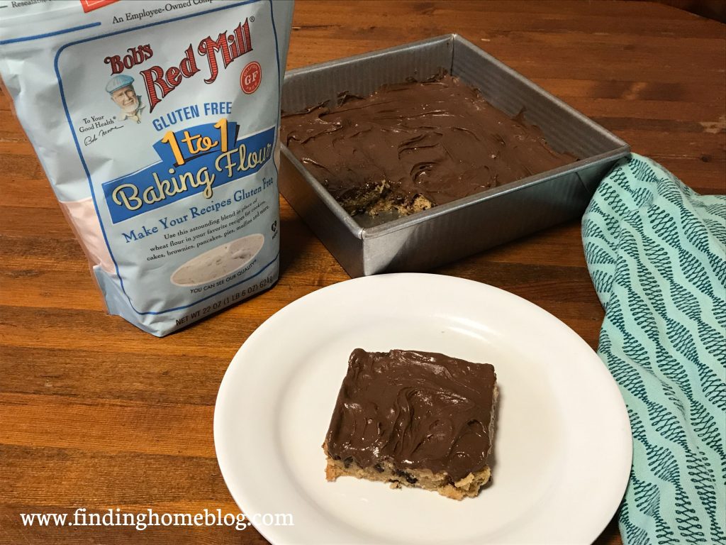 In the foreground, a peanut butter chocolate chip bar with chocolate frosting on a plate. A cloth napkin is to the right. The pan of bars is in the background. Off to the left is a package of Bob's Red Mill 1-to-1 gluten free flour.