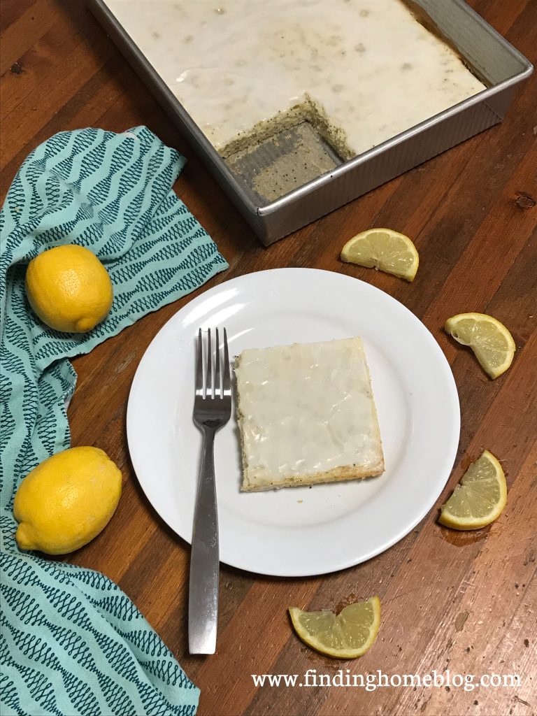 A piece of lemon cake on a plate, surrounded by lemon slices, whole lemons, a cloth napkin, and the pan of cake. 
