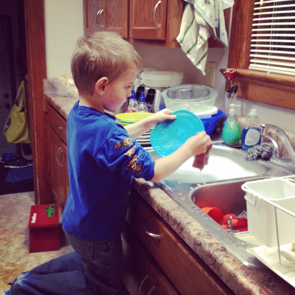 A young boy washing a plate at a sink full of soapy water.