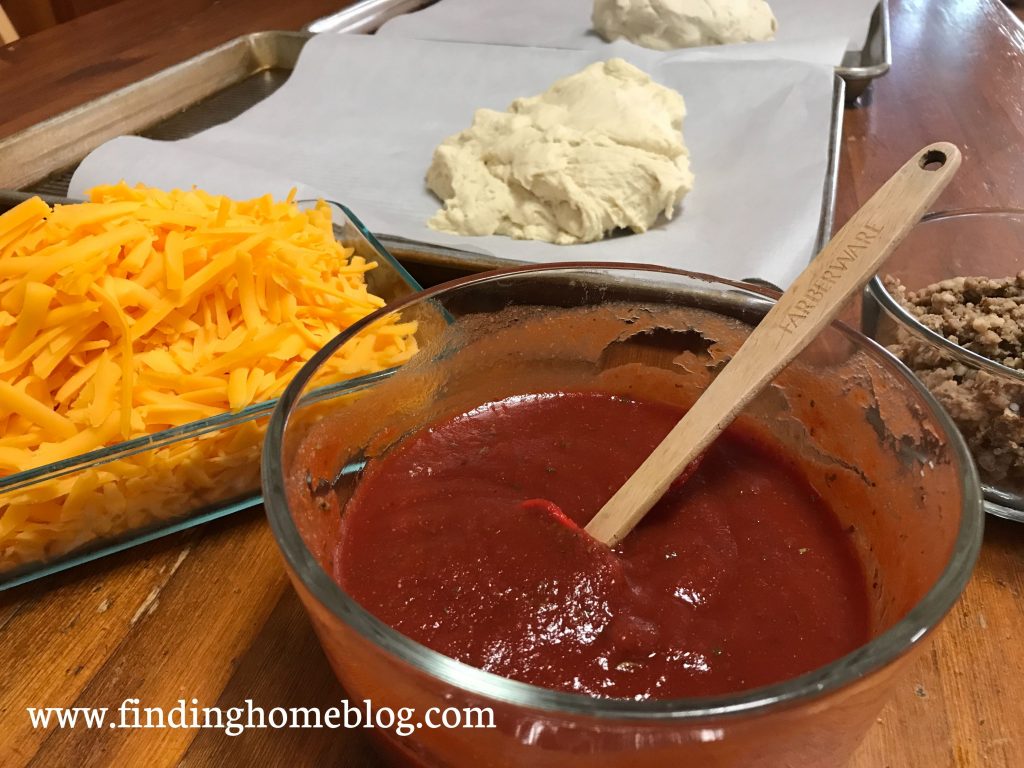 A close up view of ingredients for making pizza, with sauce and cheese in the front, and dough in the background.