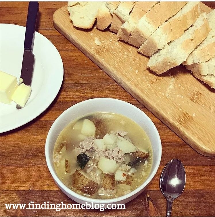 A bowl of soup with sausage, potatoes, and spinach in the foreground, with a spoon next to it. Sliced bread on a cutting board, and a plate with butter and a knife in the background.