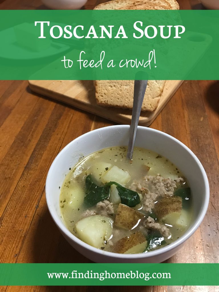 A bowl of soup with sausage, potatoes, and spinach in broth, with a spoon sticking out of it in the foreground. A sliced loaf of bread in the background.