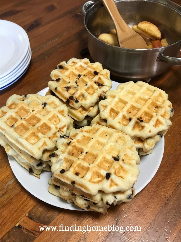 A plate with four stacks of chocolate chip waffles in the front, a pot with cinnamon apples in the back