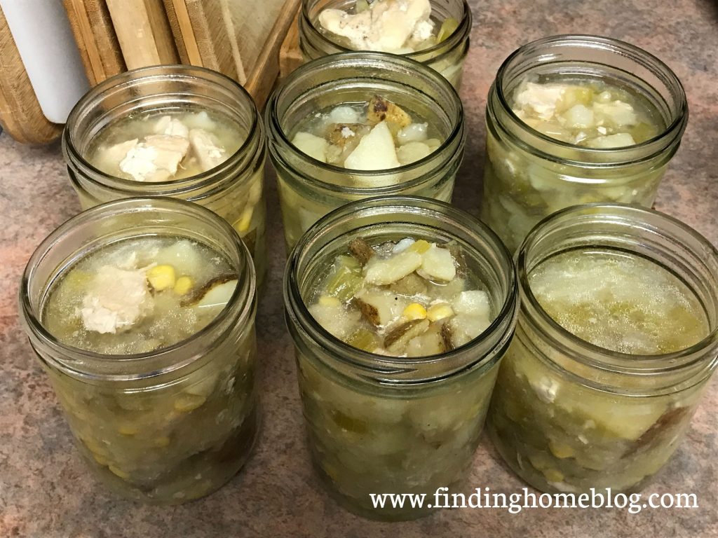 Seven jars of soup with chicken, corn, and potatoes, sitting on a counter next to some cutting boards.