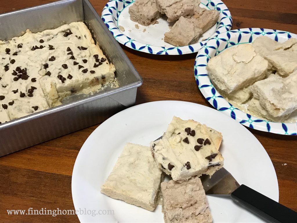 A plate with three types of shortbread bars and a small spatula. On the left, a metal pan with chocolate chip shortbread bars inside. Two paper plates with spiced and original shortbread bars in the background.