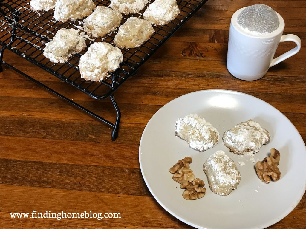 Several powdered-sugar-coated cookies on a cooling rack, as well as three on a plate with some walnuts. A powdered sugar shaker in the background.