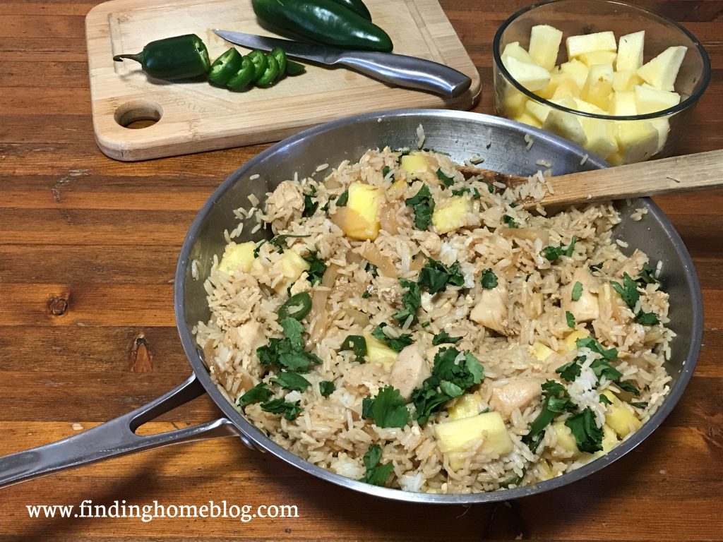 In the foreground, a large skillet with fried rice, jalapeños, and pineapple in it, and a wooden spoon. In the background, a cutting board with a knife and sliced jalapeños, and a glass dish holding chopped pineapple.