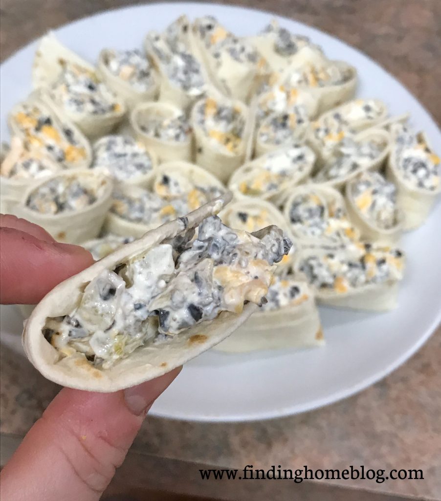 A close up on one pinwheel held between two fingers, with a plate of pinwheels - an appetizer made from tortillas rolled around a cream cheese filling - in the background