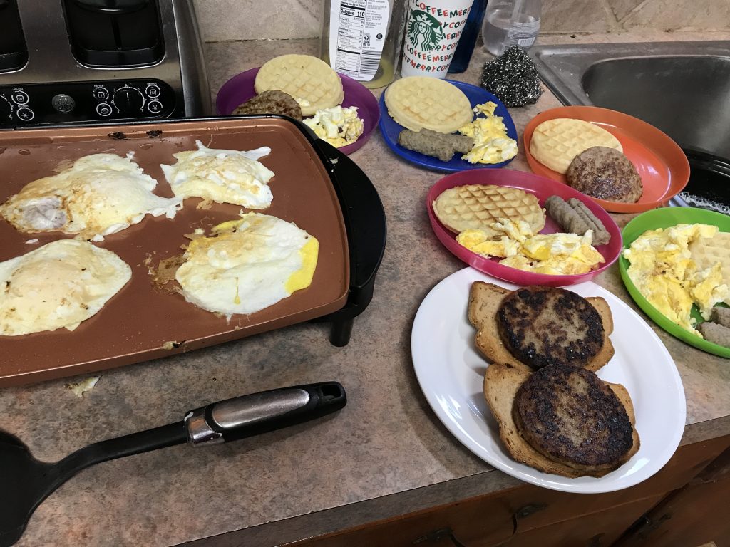 Several plates of breakfast foods: sausage, eggs, waffles, and toast, next to a griddle with more eggs cooking. 