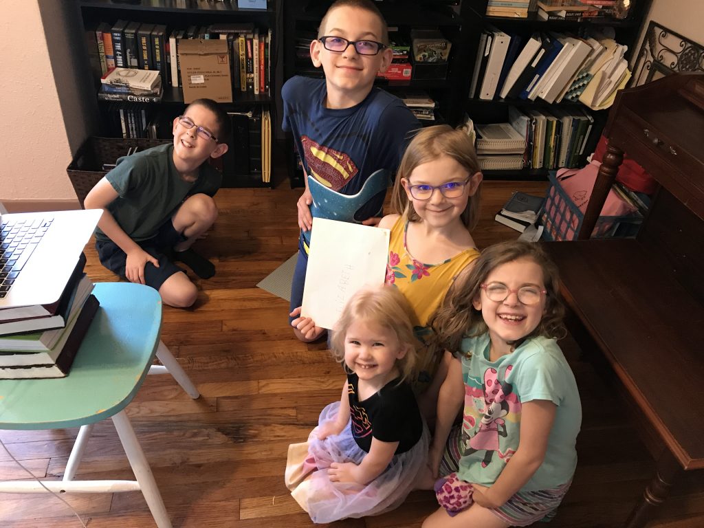 Five children gathered on the floor near a computer, propped up on books.