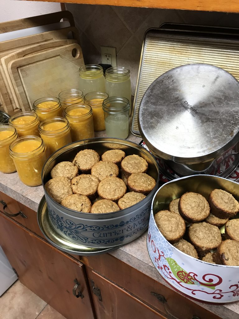 Glass jars full of a butternut soup and broth, two containers of muffins. Some clean dishes in the background.