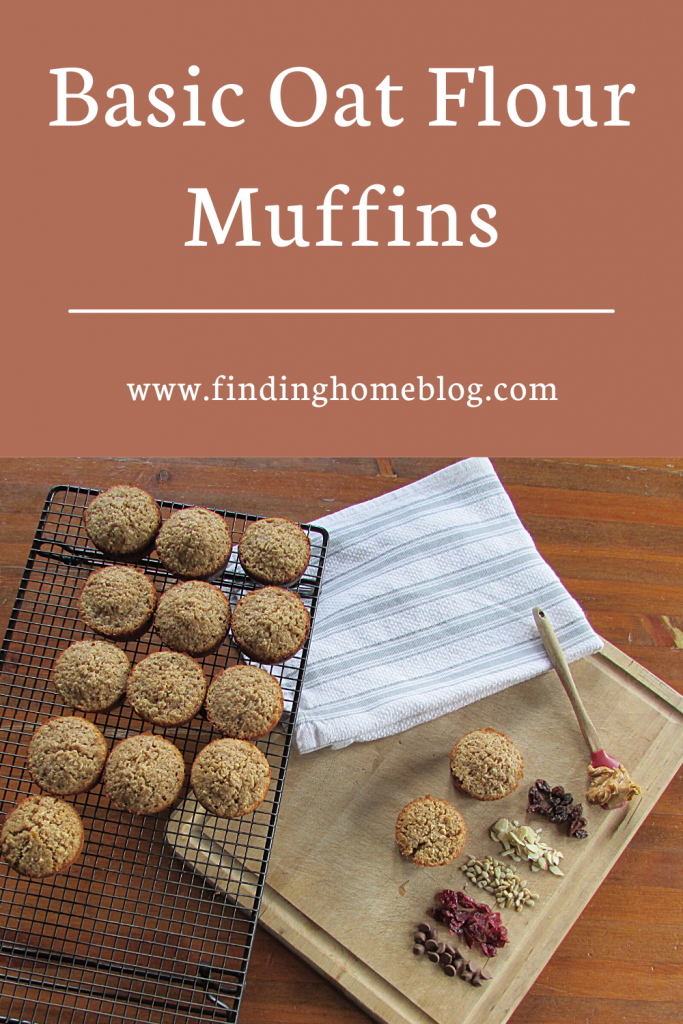 2 muffins on a wooden cutting board with a variety of toppings piled nearby: chocolate chips, dried cranberries, sunflower seeds, almonds, raisins, and a spoon with peanut butter. A cooling rack with more muffins nearby. A banner at the top of the picture reads "Basic Oat Flour Muffins"