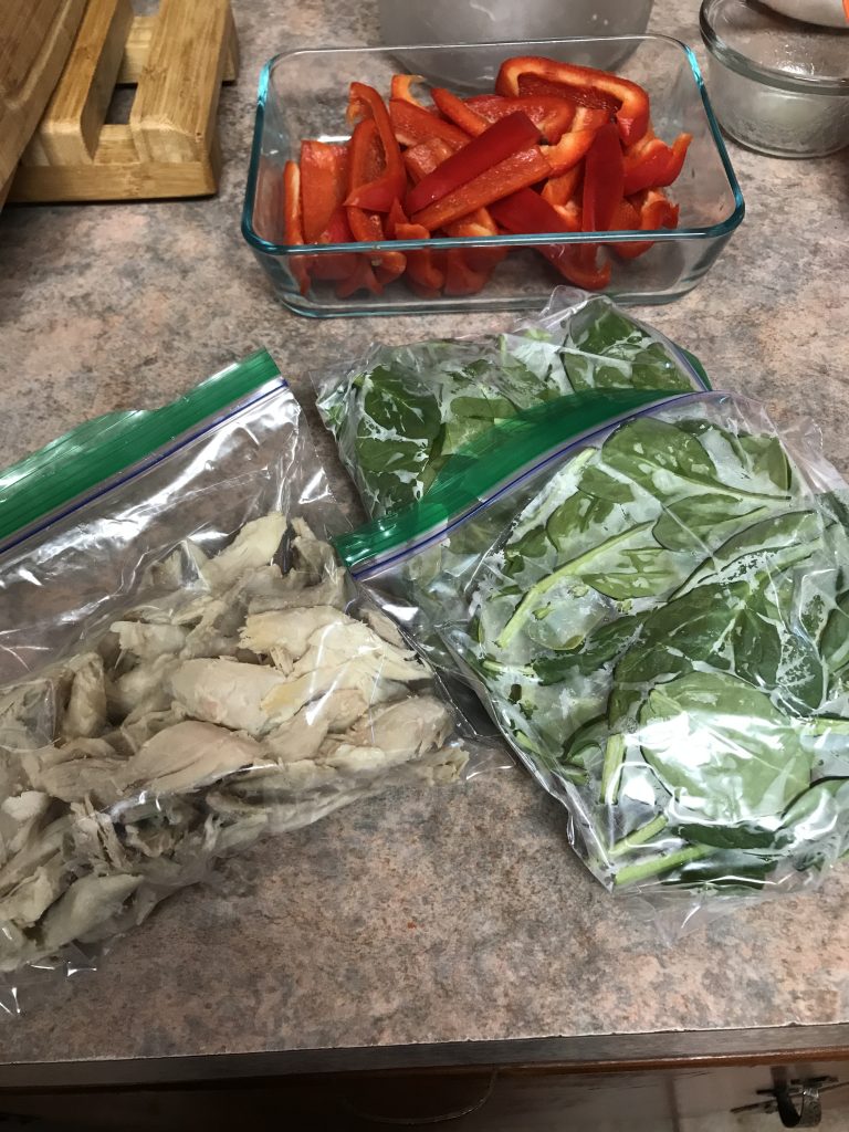 Chopped cooked chicken in a ziploc bag, 2 bags of chopped spinach nearby. A container of sliced red peppers in the background.