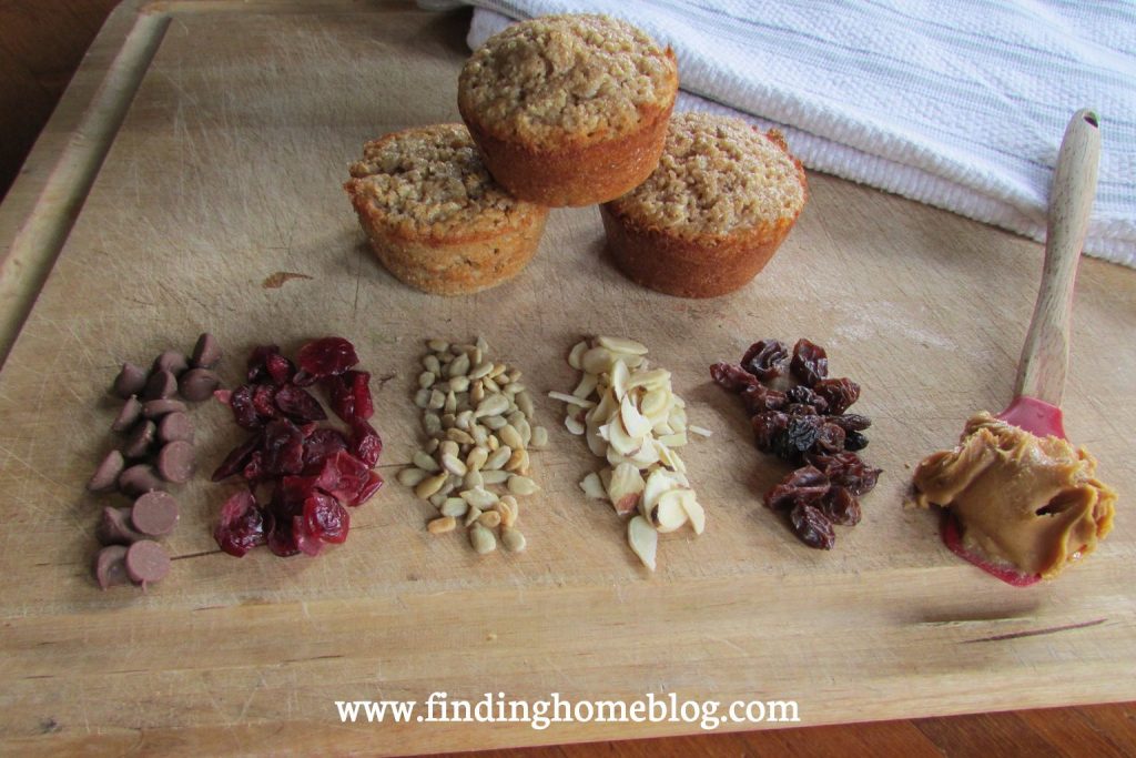 3 muffins on a wooden cutting board with a variety of toppings piled nearby: chocolate chips, dried cranberries, sunflower seeds, almonds, raisins, and a spoon with peanut butter. 