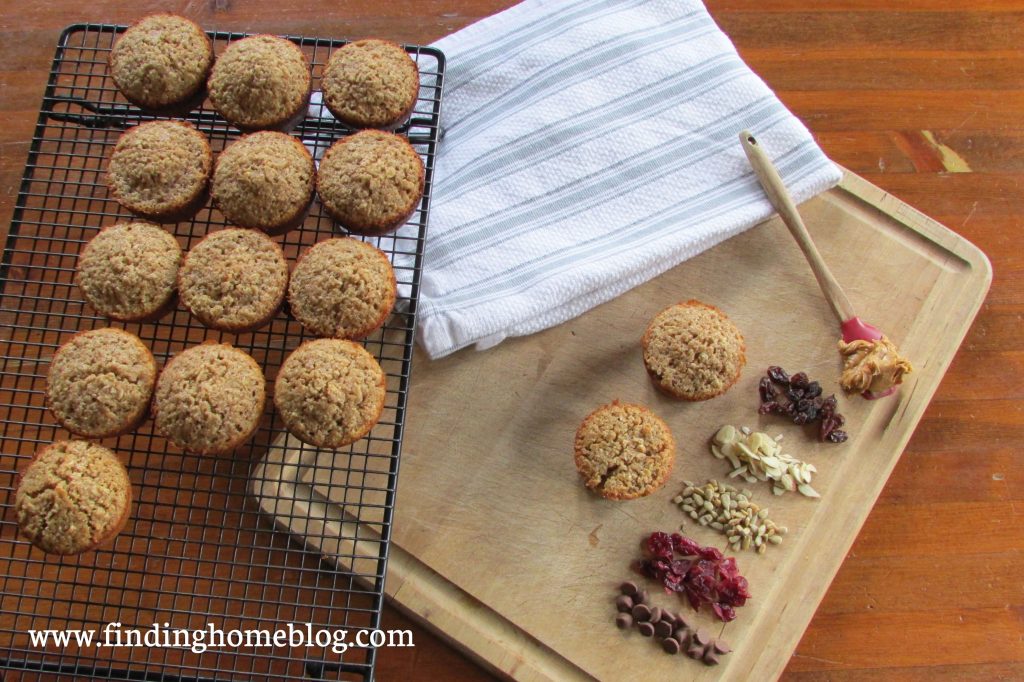 2 muffins on a wooden cutting board with a variety of toppings piled nearby: chocolate chips, dried cranberries, sunflower seeds, almonds, raisins, and a spoon with peanut butter. A cooling rack with more muffins nearby. 