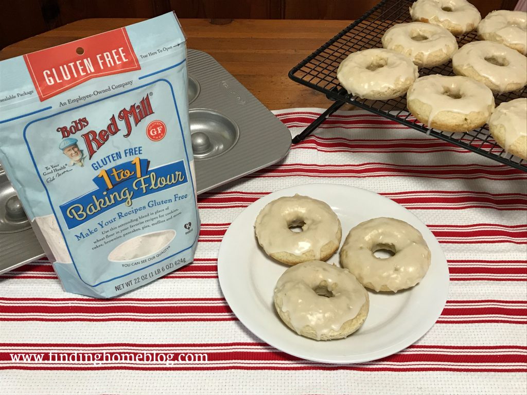 A plate with three glazed cake donuts on it in the center, with a cooling rack holding more donuts off to the right. A package of Bob's Red Mill gluten free 1-to-1 baking flour and a donut pan off to the left.