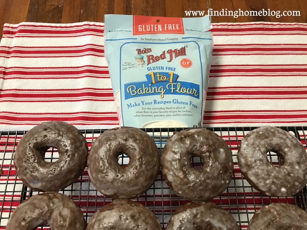 An overhead shot of four chocolate glazed donuts on a cooling rack, with a package of Bob's Red Mill 1-to-1 Gluten Free Baking Flour in the background.