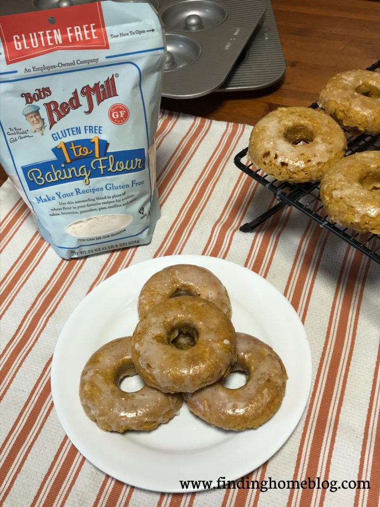 A plate of pumpkin glazed donuts in the center. Off to the left is a package of Bob's Red Mill gluten free 1-to-1 baking flour. Off to the right is a rack with more donuts on it. In the background are two donut baking pans.