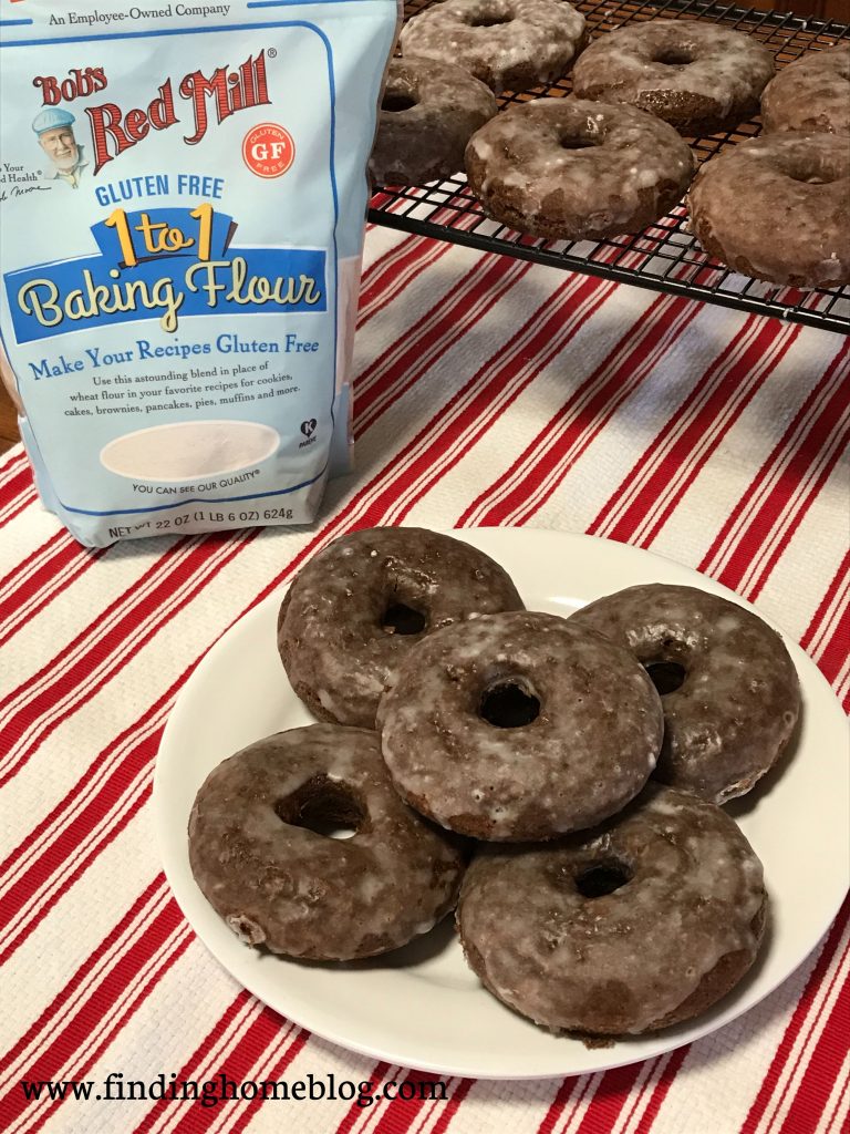 A plate of several glazed chocolate donuts in the foreground, with more on a cooling rack in the background, along with a package of Bob's Red Mill Gluten Free 1-to-1 Baking Flour.