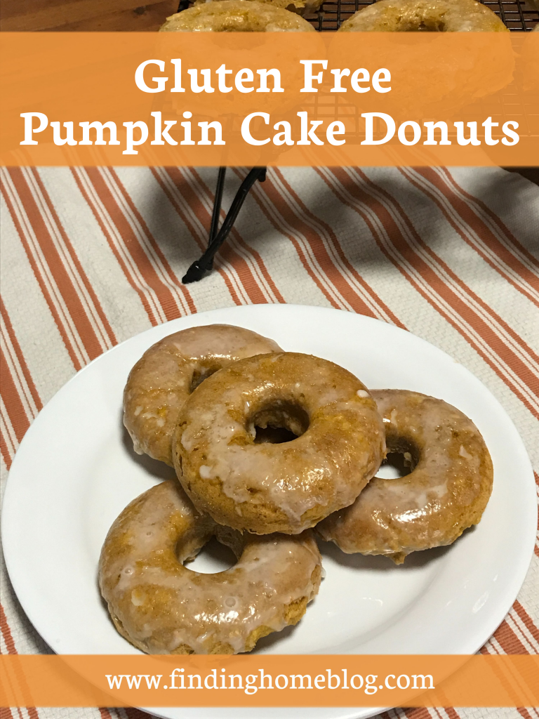 A plate of pumpkin glazed donuts sitting on a cloth, with more donuts on a rack in the background. A banner reads "Gluten Free Pumpkin Cake Donuts"
