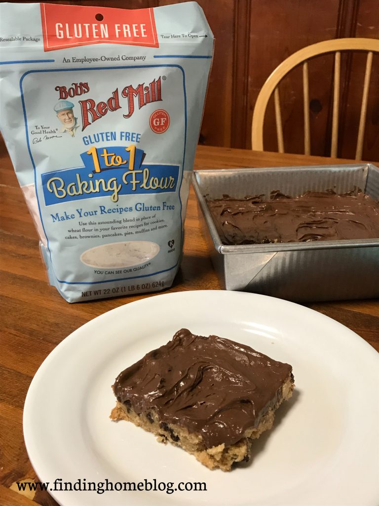 In the foreground, a peanut butter chocolate chip bar with chocolate frosting on a plate. The pan of bars is in the background. Off to the left is a package of Bob's Red Mill 1-to-1 gluten free flour.