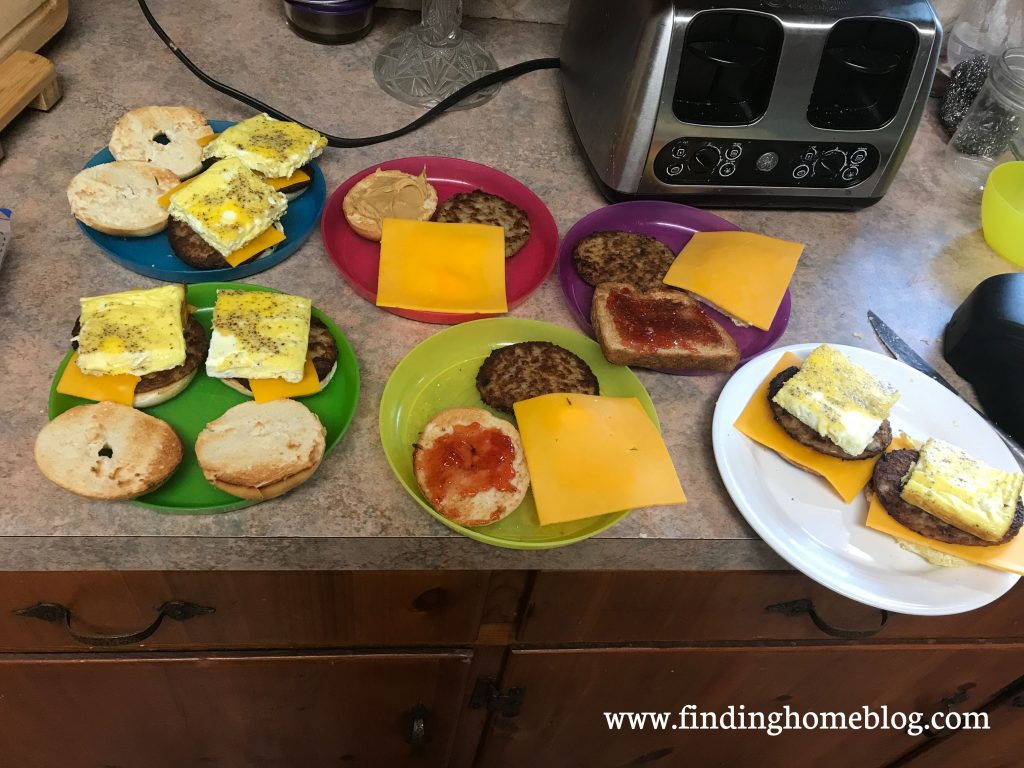 Several plates with breakfast sandwiches in various states of assembly (eggs, cheese, sausage patties, on bagels). A toaster is nearby.