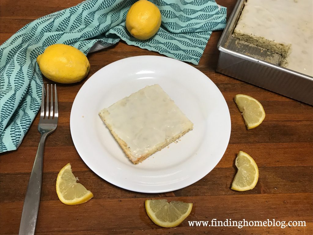 A piece of lemon cake on a plate, surrounded by lemon slices, whole lemons, a cloth napkin, and the pan of cake. 