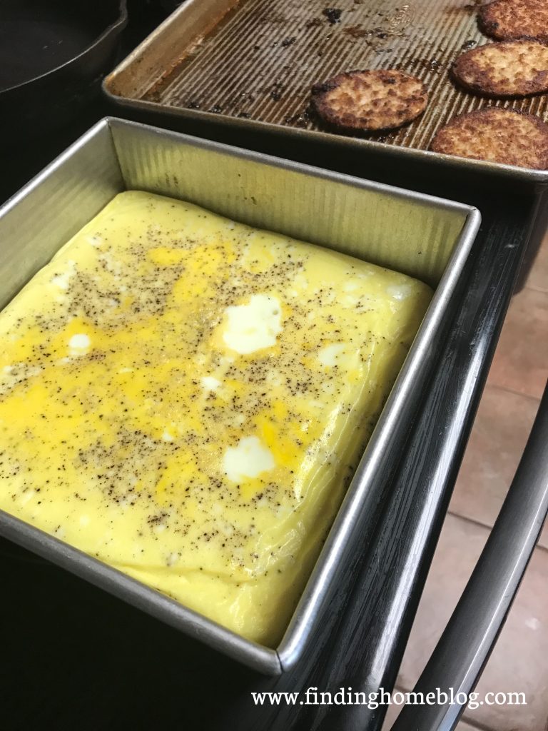 In the foreground, a pan with baked scrambled eggs. In the background, a baking sheet with cooked sausage patties.