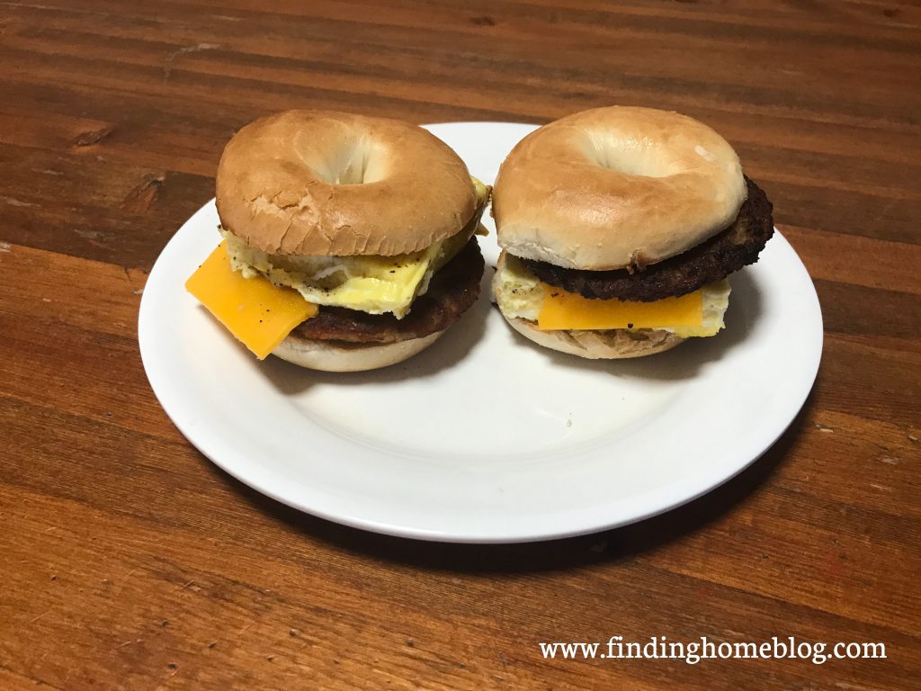 Two breakfast sandwiches on a plate, made up of bagels, sausage patties, cheese slices, and scrambled eggs.